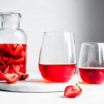Two glasses of strawberry wine on a marble tray next to a large pitcher of strawberries soaking in white wine