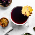 Moon tea in a tea mug shot from overhead and garnished with fresh sliced ginger. Medicinal herbs (red raspberry leaf, ginkgo and mint are in small bowls surrounding the tea).