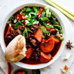 Bowlful of Bo Kho (Vietnamese Beef Stew) garnished with green onions, mint Thai basil, Thai chilies, and a baguette. Herbs and spices in background.