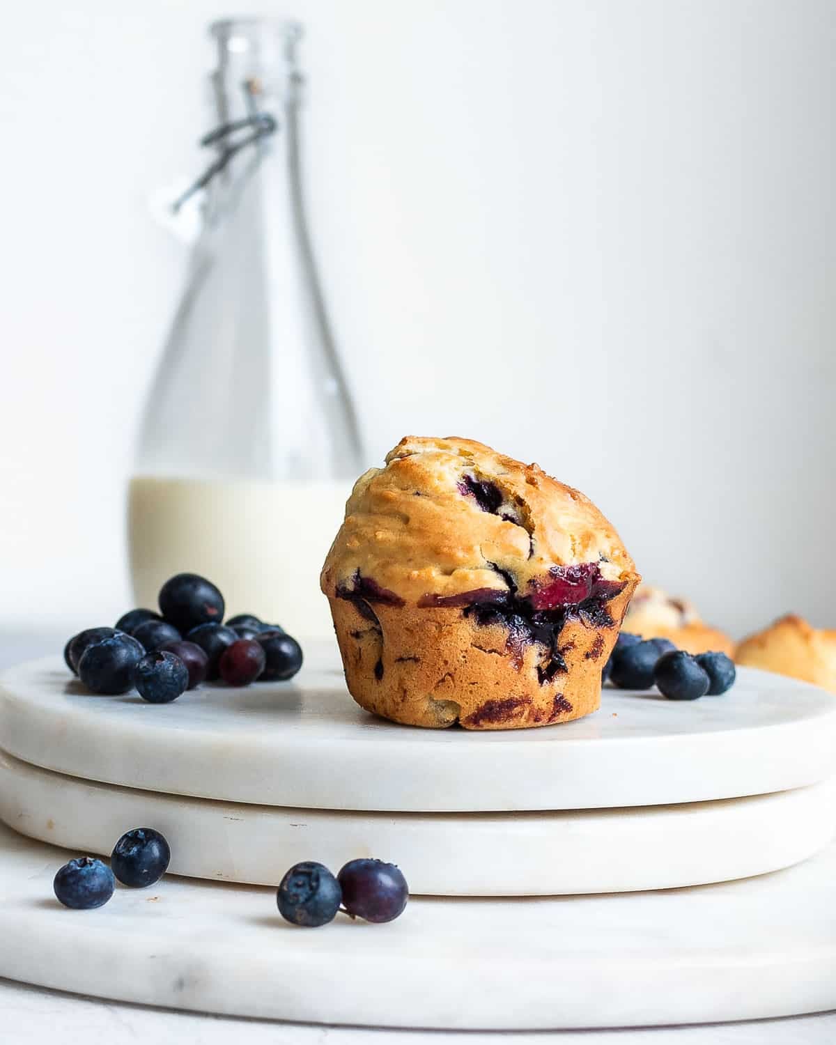 Einkorn blueberry muffin on marble surface scattered with blueberries. Milk bottle in background.