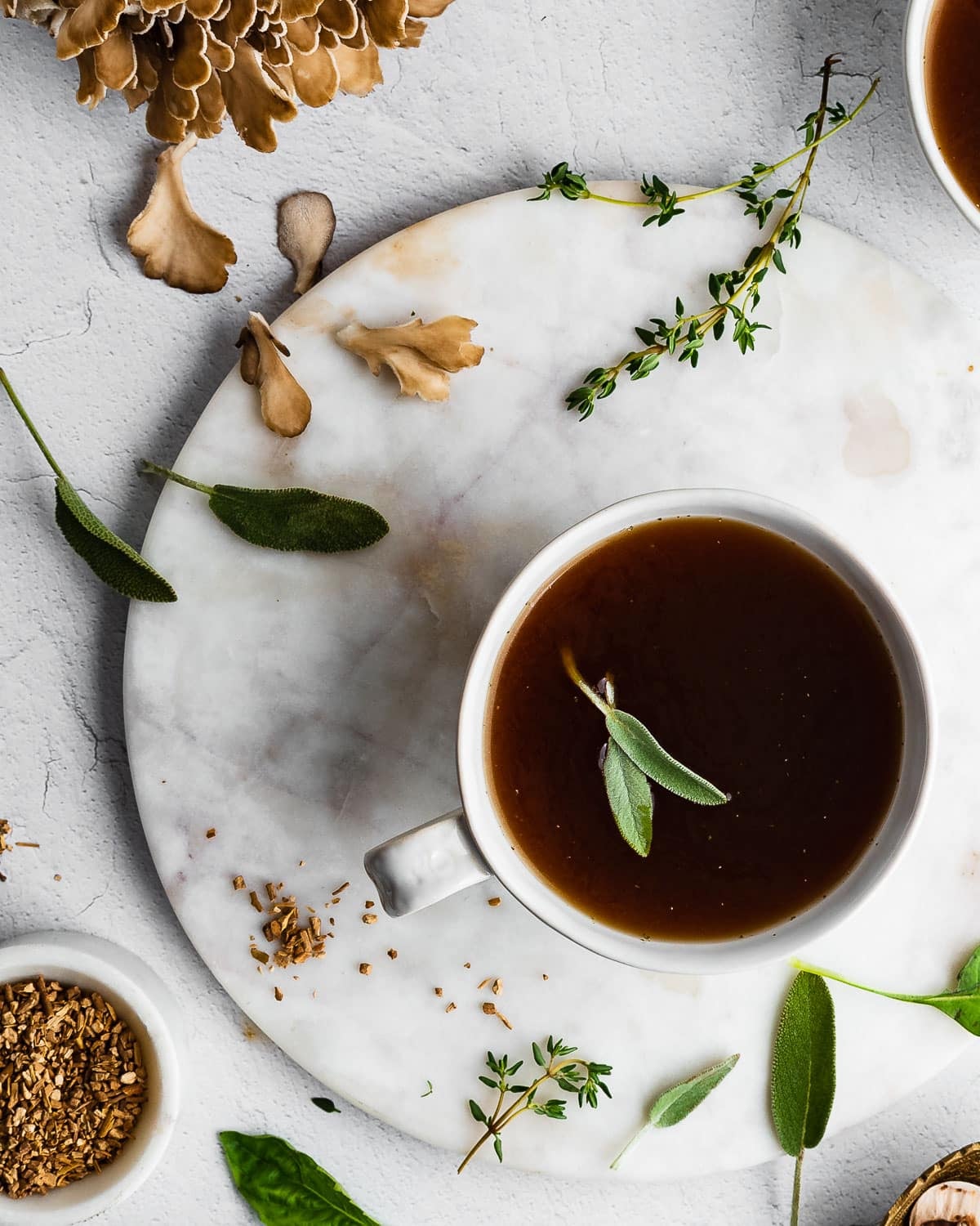 Herbal sipping broth in a white mug, herbs and mushrooms in the background