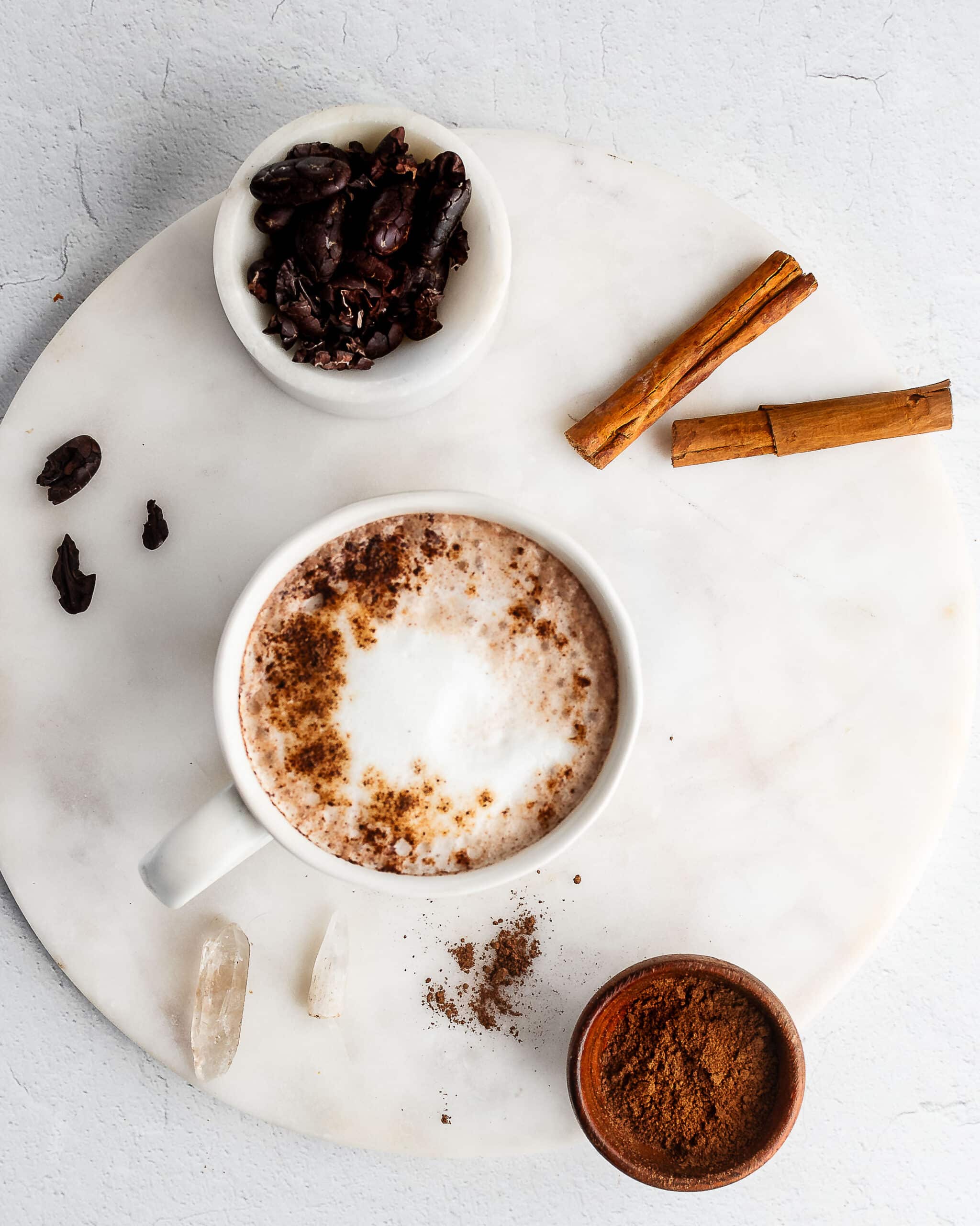 Chocolate tea latté in a white cup garnished with tea powder, cacao beans, and cinnamon sticks