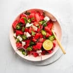 watermelon feta salad in a pink bowl garnished with lime, herbs, jalapeno and red onion