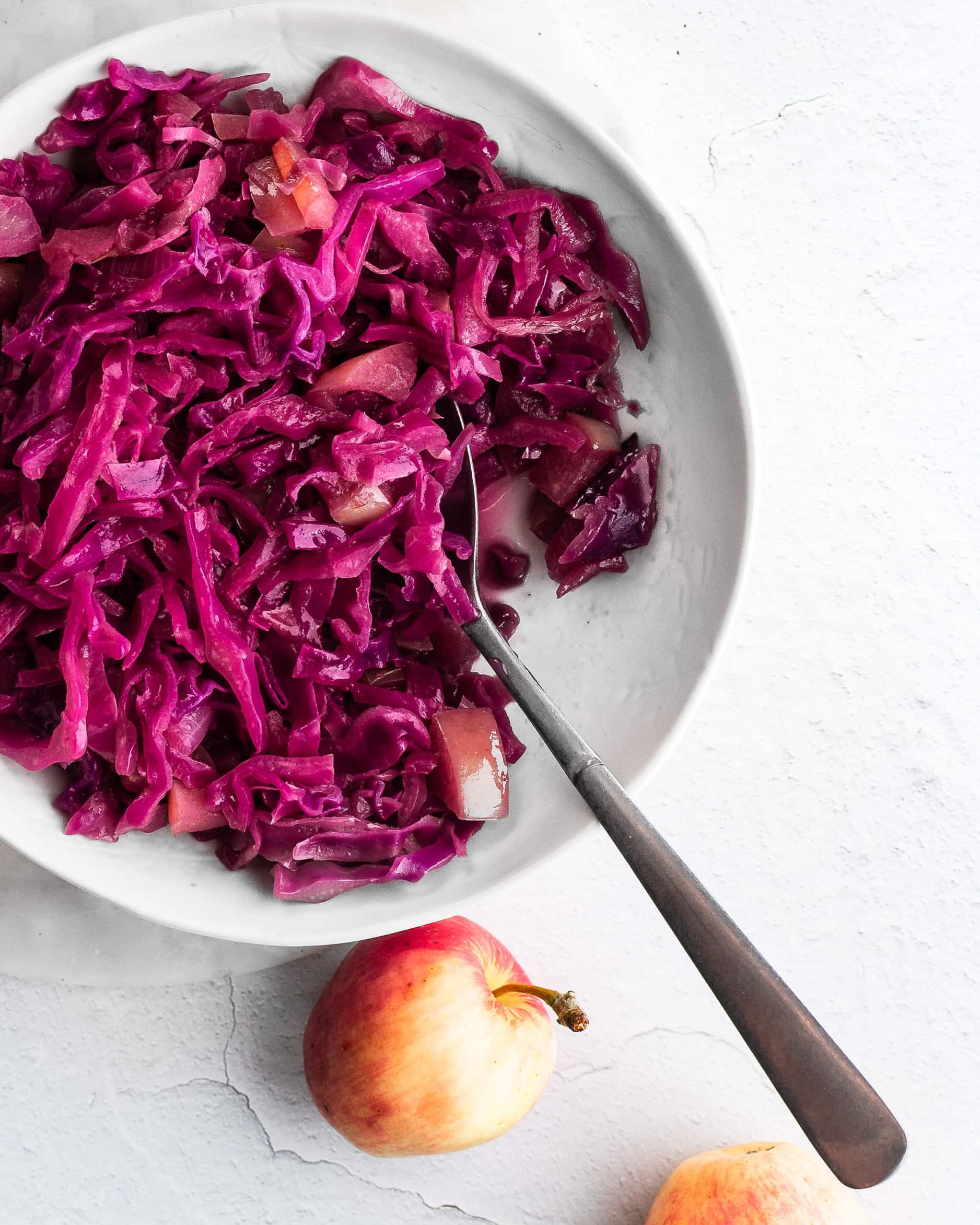 cider-braised cabbage in a white bowl, apples in background
